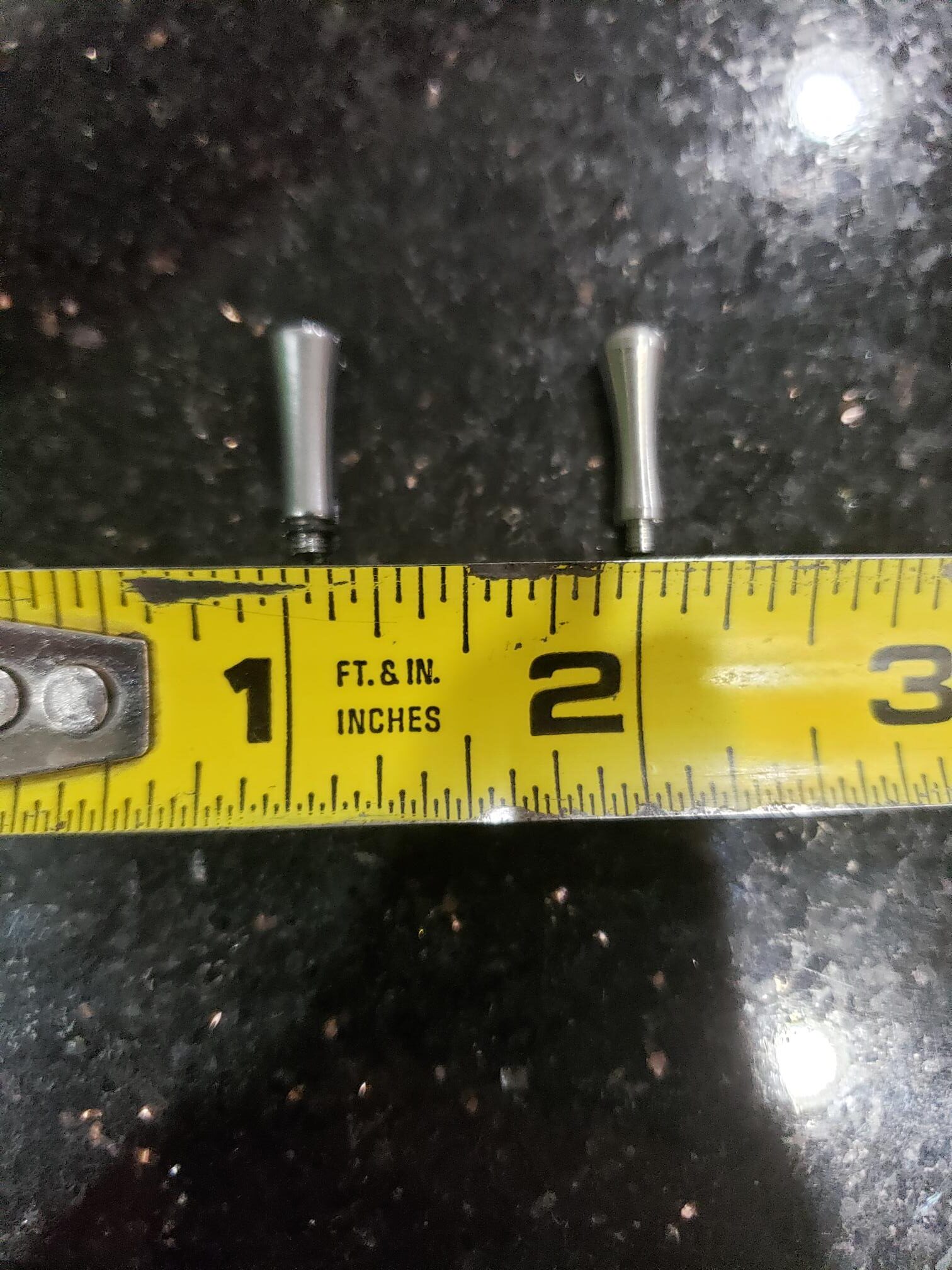 Two Tangs / Finger Rests, each about 1.5 inches long, are placed above a tape measure on a black speckled surface.