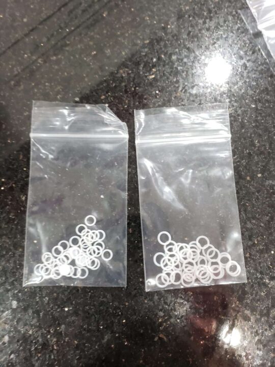 Two small transparent plastic bags containing multiple white circular washers are placed on a dark, reflective surface.